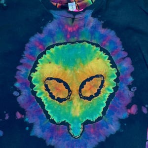 May include: A black t-shirt featuring a central tie-dye design in vibrant shades of yellow, green, blue, and purple. The pattern forms a distinct alien head shape with dark outlines defining the eyes and facial structure against the dark fabric.