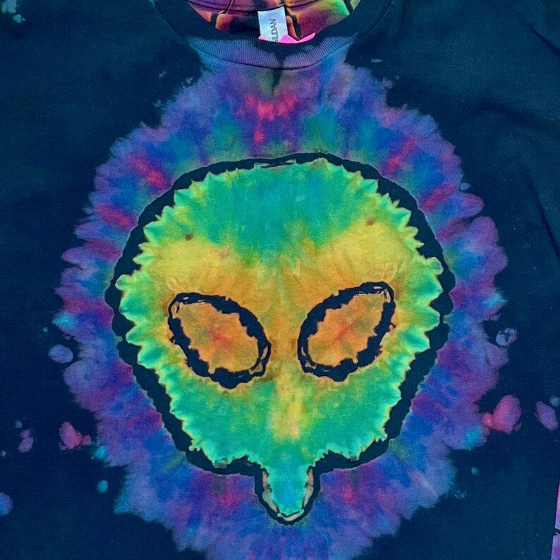 May include: A black t-shirt featuring a central tie-dye design in vibrant shades of yellow, green, blue, and purple. The pattern forms a distinct alien head shape with dark outlines defining the eyes and facial structure against the dark fabric.