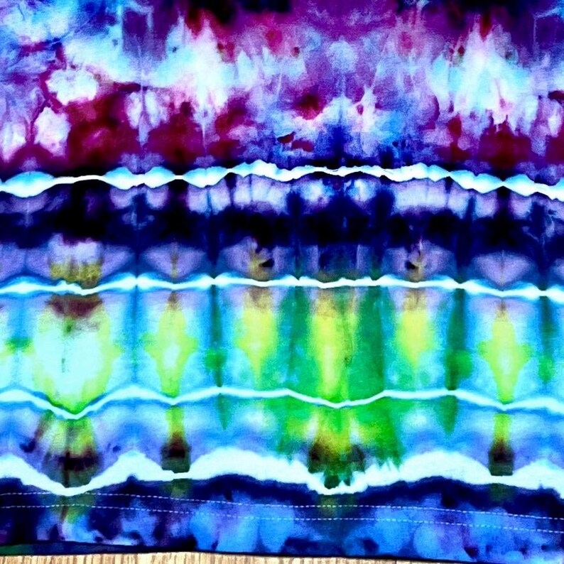 May include: A vibrant tie-dye textile featuring a blend of colors. The design showcases bands of purple, blue, green, and white, creating a visually dynamic pattern. The fabric appears soft and flowing, with a handmade aesthetic.