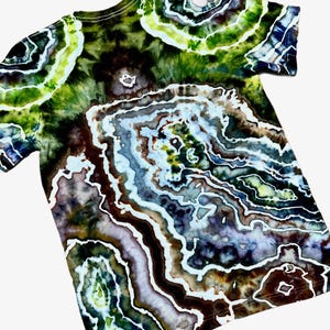 May include: A short-sleeve t-shirt featuring a complex tie-dye pattern that mimics the concentric, layered appearance of a geode. The design incorporates deep shades of forest green, dark brown, black, and soft lavender with white outlines.