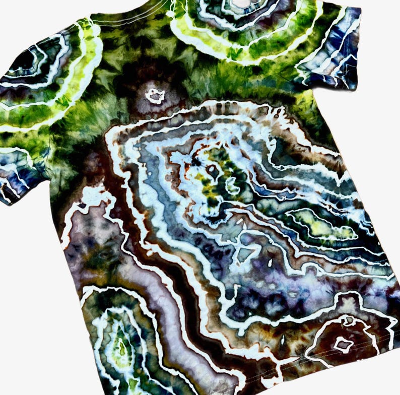 May include: A short-sleeve t-shirt featuring a complex tie-dye pattern that mimics the concentric, layered appearance of a geode. The design incorporates deep shades of forest green, dark brown, black, and soft lavender with white outlines.
