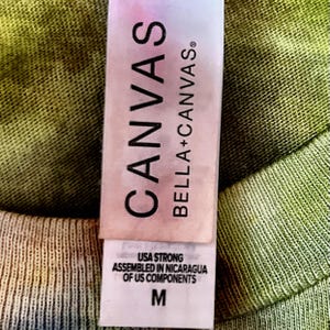May include: A white clothing tag on a tie-dye olive green fabric. The tag features the text CANVAS BELLA+CANVAS in black, followed by USA STRONG ASSEMBLED IN NICARAGUA OF US COMPONENTS and the size letter M printed in a clear, bold font.