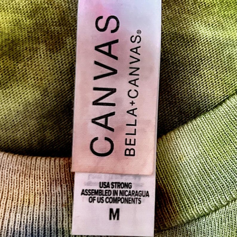 May include: A white clothing tag on a tie-dye olive green fabric. The tag features the text CANVAS BELLA+CANVAS in black, followed by USA STRONG ASSEMBLED IN NICARAGUA OF US COMPONENTS and the size letter M printed in a clear, bold font.