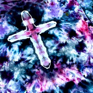 May include: A fabric garment featuring a tie-dye pattern in shades of deep blue, purple, and black. A prominent, un-dyed white cross shape is centered on the fabric, with soft, blurred edges and subtle pinkish-purple hues within the white area.