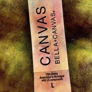 May include: A rectangular clothing tag with a tie-dye pattern in shades of pink, blue, and yellow. The text reads CANVAS BELLA+CANVAS, USA Strong, Assembled in Nicaragua with US components, and the size letter L is printed at the bottom.