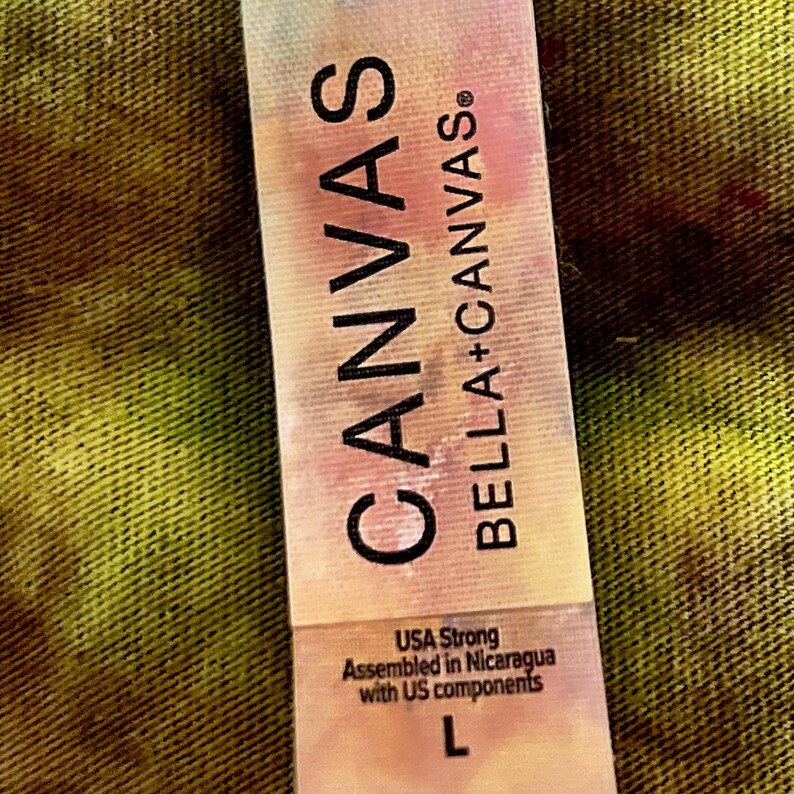 May include: A rectangular clothing tag with a tie-dye pattern in shades of pink, blue, and yellow. The text reads CANVAS BELLA+CANVAS, USA Strong, Assembled in Nicaragua with US components, and the size letter L is printed at the bottom.