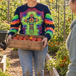 May include: A long-sleeved navy blue shirt featuring a vibrant tie-dye cross pattern in shades of red, orange, yellow, green, and purple, paired with light blue denim jeans. The shirt has a crew neckline and ribbed cuffs on the sleeves.