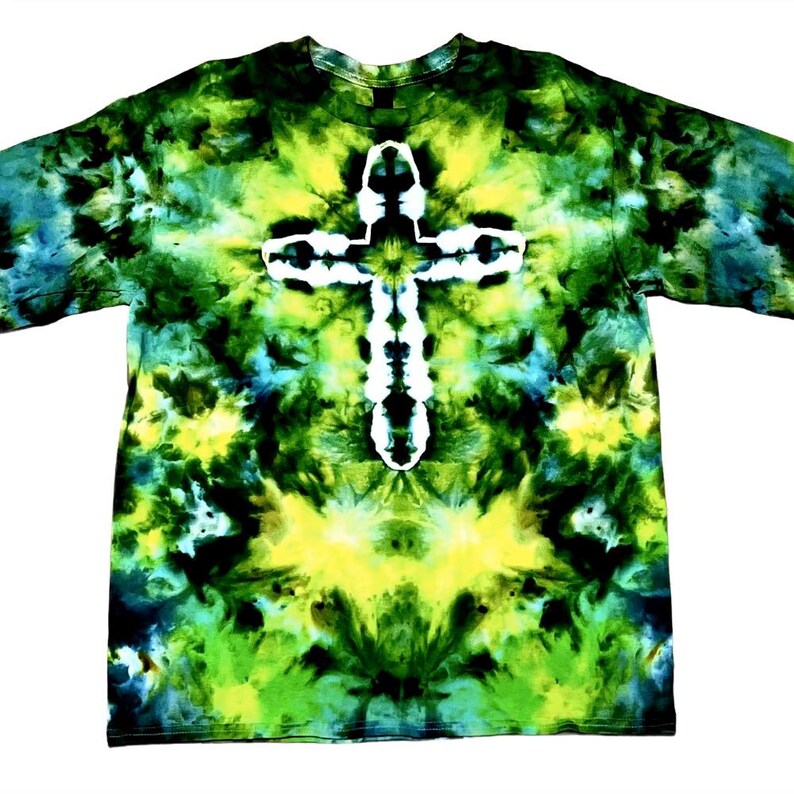 May include: A short-sleeve t-shirt featuring a tie-dye pattern in shades of forest green, lime green, black, and teal. The center of the shirt displays a prominent white cross shape created through the tie-dye process against the dark background.