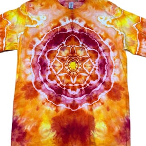 Puede incluir: Una camiseta de manga corta con un patr&oacute;n tie-dye en tonos naranja, amarillo y rojo intenso. El centro de la camiseta muestra un dise&ntilde;o circular y sim&eacute;trico tipo mandala con un n&uacute;cleo amarillo brillante rodeado de anillos conc&eacute;ntricos.