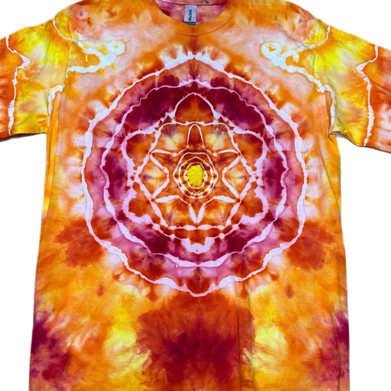Puede incluir: Una camiseta de manga corta con un patr&oacute;n tie-dye en tonos naranja, amarillo y rojo intenso. El centro de la camiseta muestra un dise&ntilde;o circular y sim&eacute;trico tipo mandala con un n&uacute;cleo amarillo brillante rodeado de anillos conc&eacute;ntricos.