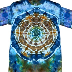 May include: A short-sleeved t-shirt featuring a tie-dye pattern with a central circular mandala design. The colours include shades of deep blue, light blue, brown, and hints of green, separated by distinct white lines throughout the fabric.