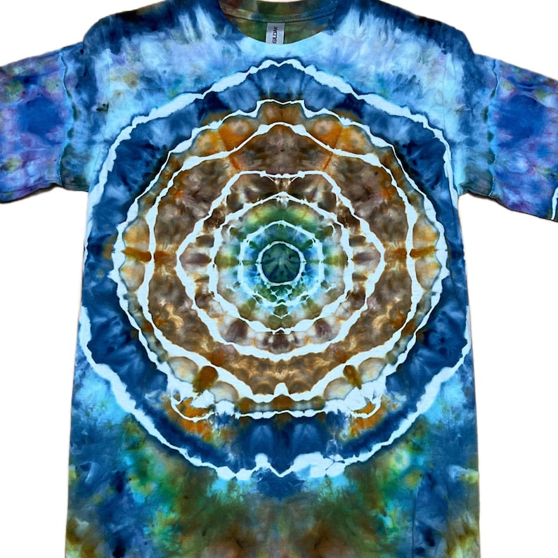 May include: A short-sleeved t-shirt featuring a tie-dye pattern with a central circular mandala design. The colours include shades of deep blue, light blue, brown, and hints of green, separated by distinct white lines throughout the fabric.