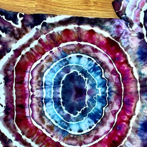Puede incluir: Una camiseta con un patr&oacute;n tie-dye circular que presenta anillos conc&eacute;ntricos de azul profundo, magenta vibrante y morado oscuro. El dise&ntilde;o incluye l&iacute;neas blancas finas e irregulares que separan los colores, creando un efecto radiante.