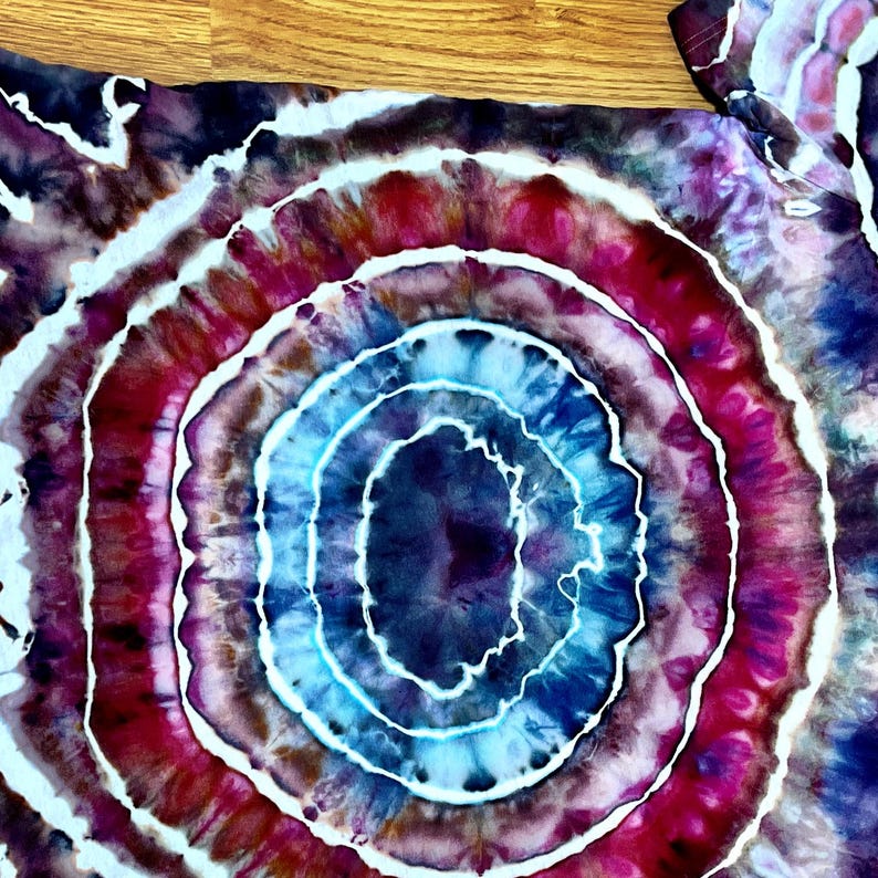 Puede incluir: Una camiseta con un patr&oacute;n tie-dye circular que presenta anillos conc&eacute;ntricos de azul profundo, magenta vibrante y morado oscuro. El dise&ntilde;o incluye l&iacute;neas blancas finas e irregulares que separan los colores, creando un efecto radiante.
