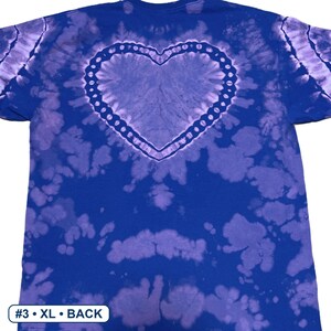 Peut inclure: Un t-shirt &agrave; manches courtes avec un motif tie-dye dans des tons bleu roi et violet clair. Le dos du t-shirt pr&eacute;sente une forme de c&oelig;ur pro&eacute;minente soulign&eacute;e par une bordure pointill&eacute;e d&eacute;corative. Le texte #3 &bull; XL &bull; BACK est visible.