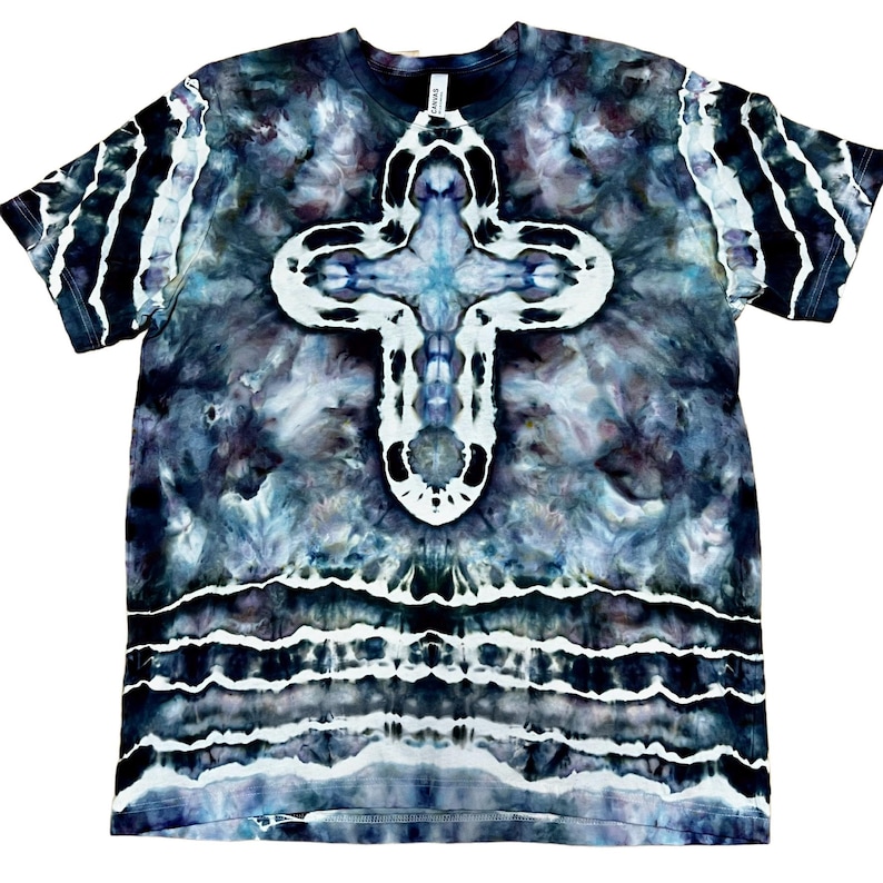 May include: A tie-dye t-shirt with a large white and blue cross design centered on the front. The shirt has a dark blue and gray background with white stripes on the sleeves and bottom hem. The shirt is a crew neck.