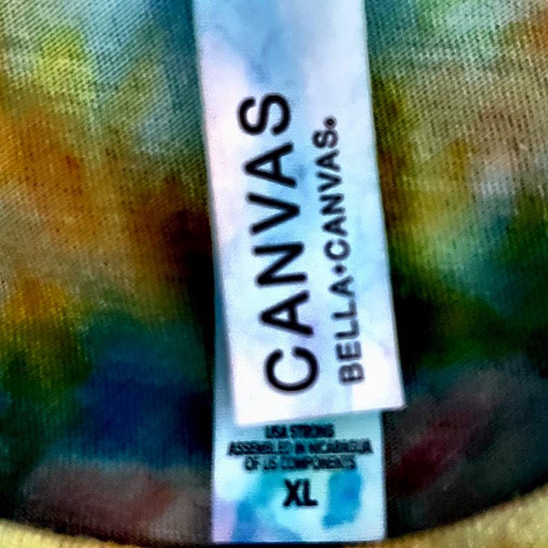 May include: A white fabric clothing tag with black text reading CANVAS BELLA+CANVAS, USA STRONG, ASSEMBLED IN NICARAGUA OF US COMPONENTS, and XL, attached to a tie-dye garment featuring blended swirls of yellow, green, blue, and orange colors.