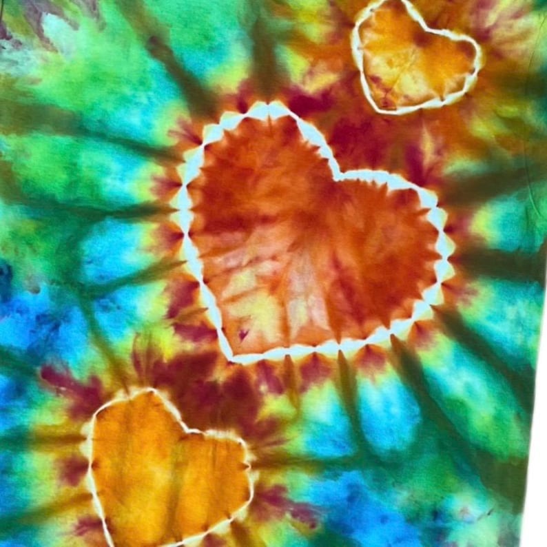 Może przedstawiać: Tkanina farbowana metodą tie-dye z trzema wyraźnymi kształtami serc w odcieniach pomarańczu i ż&oacute;łci. Serca są obwiedzione białymi krawędziami i umieszczone na żywym tle z mieszanymi wzorami tie-dye w kolorach zielonym, niebieskim i czerwonym.