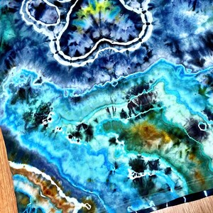 May include: A tie-dye shirt in shades of blue, teal, and yellow. The fabric features a marbled pattern with white outlines, creating a unique, artistic design. The shirt's design resembles a landscape with water and clouds.