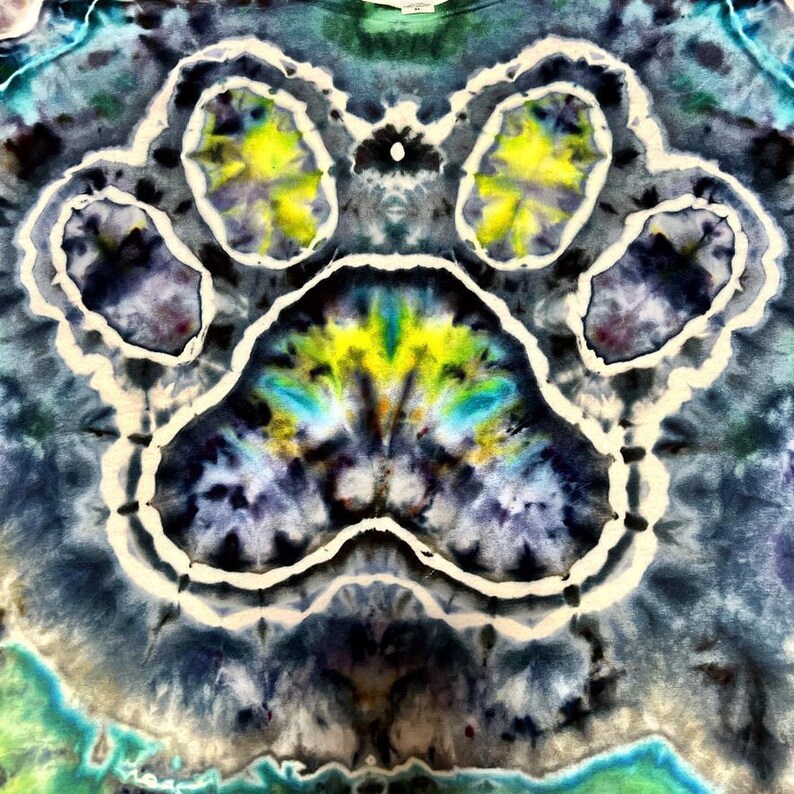 May include: Tie-dye t-shirt featuring a large paw print design. The paw print is outlined in white and filled with yellow, blue, and green hues. The background is a blend of blues, greens, and purples, creating a vibrant, colorful pattern.
