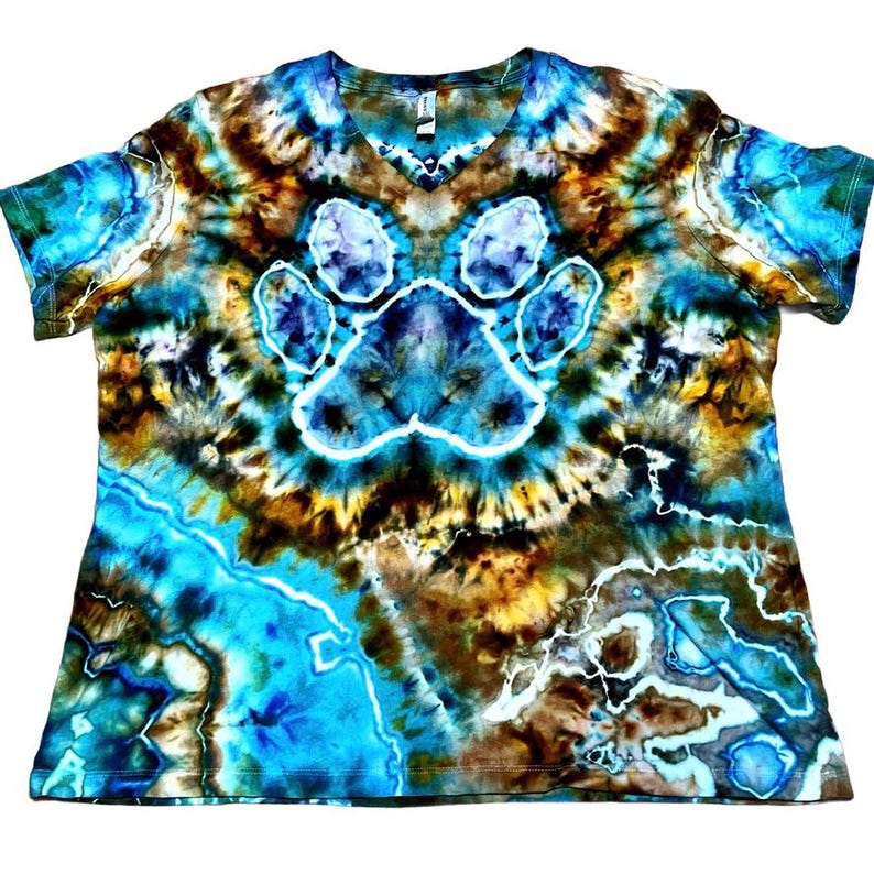 May include: A short-sleeve V-neck t-shirt featuring a tie-dye pattern in shades of bright blue, dark brown, and white. The center of the shirt displays a prominent paw print graphic outlined in white, surrounded by swirling, marbled color effects.