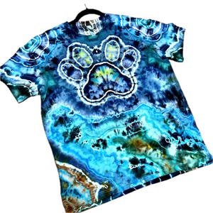 May include: A tie-dye t-shirt in shades of blue, teal, and brown, featuring a large white outline of a paw print in the center. The shirt has a crew neck and short sleeves, hanging on a black hanger.