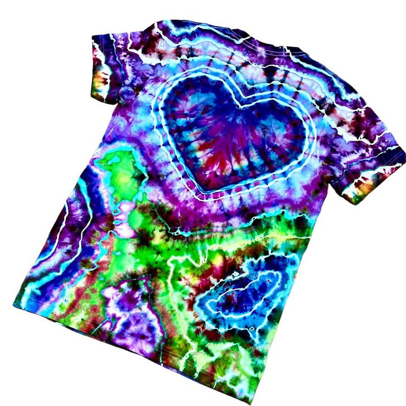 May include: A colourful tie-dye t-shirt with a heart design at the centre. The shirt displays a vibrant mix of colours, including blue, purple, green, and yellow. The heart is outlined in white, and the tie-dye pattern creates a swirling effect.