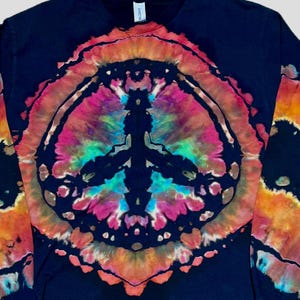 May include: A black long-sleeve shirt featuring a large, circular tie-dye peace sign graphic on the front. The design incorporates vibrant shades of pink, orange, yellow, and turquoise against the dark fabric, creating a bold, colorful pattern.
