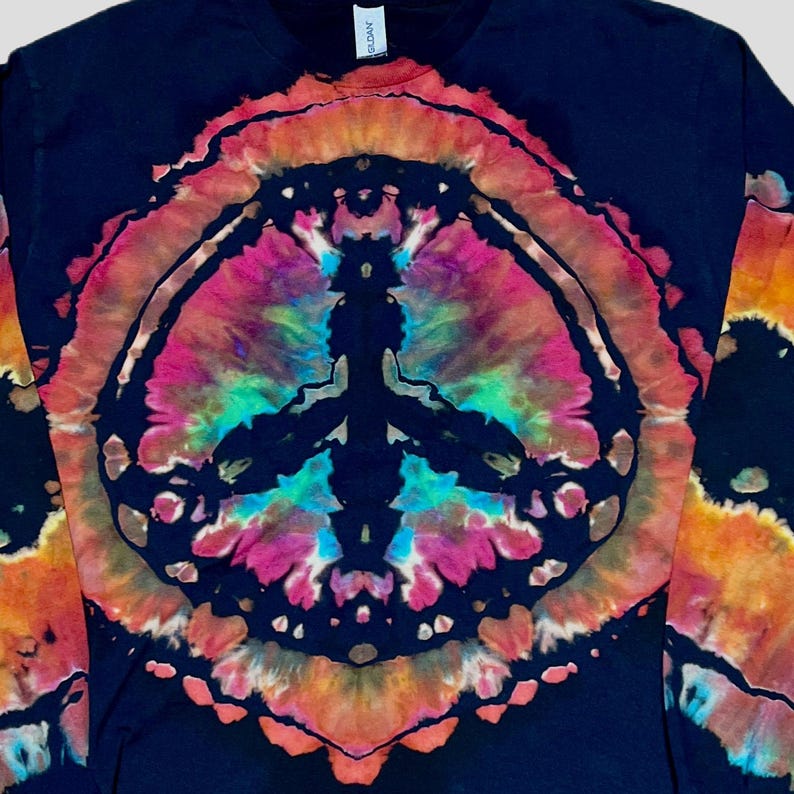 May include: A black long-sleeve shirt featuring a large, circular tie-dye peace sign graphic on the front. The design incorporates vibrant shades of pink, orange, yellow, and turquoise against the dark fabric, creating a bold, colorful pattern.