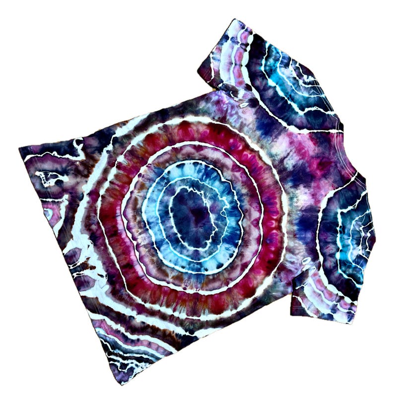 Puede incluir: Una camiseta de manga corta con un patr&oacute;n tie-dye circular de anillos conc&eacute;ntricos en azul profundo, magenta brillante y blanco. El dise&ntilde;o irradia desde el centro, creando un efecto de geoda en toda la tela con acentos de color &iacute;ndigo oscuro.