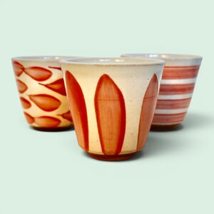 Peut inclure: Trois tasses en céramique avec une base de couleur crème et des motifs peints à la main de couleur terre cuite. La tasse avant présente des rayures verticales, celle de gauche un motif de petites feuilles et celle de droite des rayures horizontales.