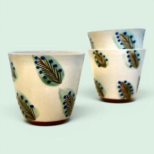 Peut inclure: Deux tasses en céramique blanc cassé ornées d'un motif répétitif de feuilles brunes semblables à des fougères, entourées d'ovales vert clair style aquarelle et de petits points bleus. Les tasses sont coniques avec une base brun foncé.