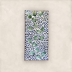 Può includere: Piatto da portata rettangolare in ceramica con sfondo bianco ricoperto di pois blu e neri. Rami di foglie verdi sono dipinti sulla superficie. Il piatto ha un bordo marrone.