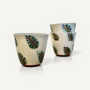 Peut inclure: Quatre tasses en céramique avec une base crème et un motif de feuilles. Les feuilles sont vertes et marron avec des accents bleus en forme de baies. Les tasses ont une forme légèrement évasée et une base marron.