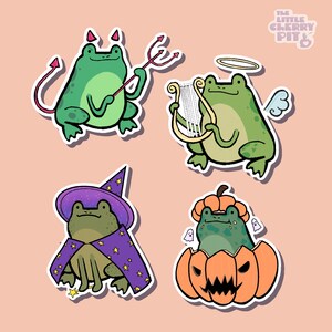 May include: Four whimsical frog stickers on a light pink background. One frog is a devil with a pitchfork, another an angel with a harp, a third a wizard, and the last a frog in a jack-o'-lantern. The Little Cherry Pit logo is in the top right corner.