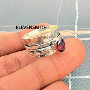 以下が含まれることがあります： ベゼルにセットされたファセットカットの楕円形レッドガーネットストーンを特徴とする、幅広のスターリングシルバー製スピナーリング。バンドはテクスチャ加工された酸化仕上げで、装飾的なエッチングのディテールと、金属表面に明確に印刷されたELEVENSMITHの文字が施されています。