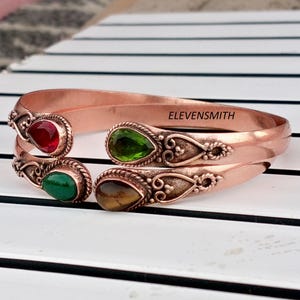 May include: A copper cuff bracelet featuring an open design with four teardrop-shaped gemstones in red, bright green, dark green, and brown. The metal ends are decorated with intricate scrollwork and the text ELEVENSMITH is engraved on the inner band.