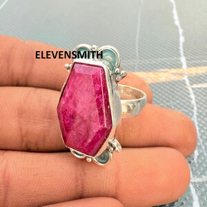 Peut inclure: Une bague en argent avec une grande pierre précieuse magenta à facettes en forme de cercueil, sertie dans une monture en métal décorative. Le mot ELEVENSMITH est imprimé en noir sur le côté gauche. L'anneau est lisse et métallique.