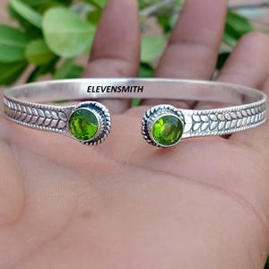 Peut inclure: Un bracelet manchette en argent avec un motif de feuilles répétitif le long de l'anneau et deux pierres précieuses péridot vertes, rondes et facettées, aux extrémités ouvertes. Le mot ELEVENSMITH est imprimé en noir sur la face interne.