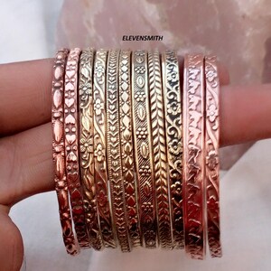 Pode incluir: Uma coleção de dez pulseiras metálicas com vários padrões em relevo, incluindo motivos florais, formas geométricas e desenhos de corações repetidos. As pulseiras exibem tons de cobre e latão com o texto ELEVENSMITH visível.