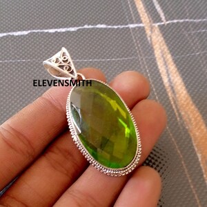 May include: A silver pendant featuring a large, oval-cut, olive-green gemstone. The pendant has a decorative silver bail and a textured silver border. The text "ELEVENSMITH" is visible on the pendant.