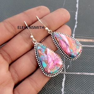 Op de afbeelding: Een paar druppelvormige oorbellen met een zilverkleurig montuur en een kleurrijke stenen inleg. De steen heeft tinten roze, turquoise en goud. De oorbellen hangen aan een eenvoudige haak.