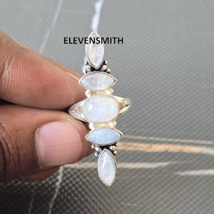 May include: A silver ring featuring five oval and marquise-cut moonstones. The ring has a vertical design with the stones set in a row. The text "ELEVENSMITH" is visible on the ring.