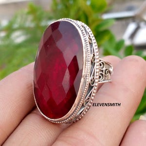Pode incluir: Um anel com uma grande pedra preciosa de cor vermelho-escura, formato oval e facetada, montada numa banda de metal prateado. O engaste apresenta filigrana intrincada e uma borda com contas. O texto ELEVENSMITH é visível na lateral.