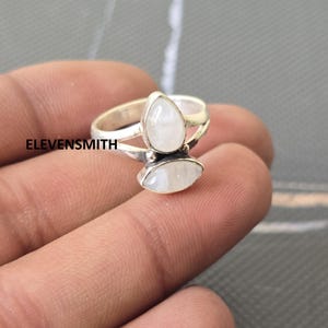 May include: A silver ring featuring two moonstone gemstones. The top stone is pear-shaped, and the bottom stone is oval. The ring is on a finger. The text "ELEVENSMITH" is visible.