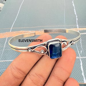 Könnte beinhalten: Ein silberfarbenes Armreif-Armband mit einem rechteckigen, tiefblauen Edelstein in der Mitte. Das Armband hat dekorative Wirbel und kleine Perlenakzente. Der Text "ELEVENSMITH" ist sichtbar.