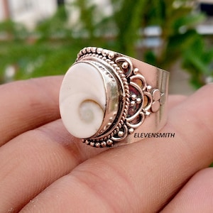 May include: A silver ring featuring a central oval-shaped white shiva eye shell with a natural spiral pattern. The metal band displays intricate decorative metalwork with beaded and filigree details. The text ELEVENSMITH is printed on the band.