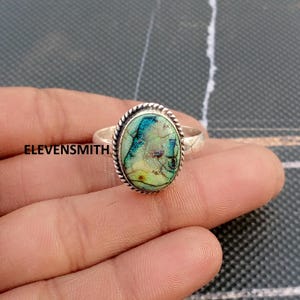 May include: A silver ring with an oval-shaped, iridescent stone. The stone has shades of blue, green, and yellow with a unique pattern. The ring band is simple, and the stone is framed by a twisted rope design. The text "ELEVENSMITH" is visible.