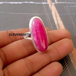 Op de afbeelding: Een zilveren ring met een grote, ovaalvormige, roze edelsteen. De edelsteen heeft een gemarmerd uiterlijk. De ring is gezet in een zilveren band. De tekst "ELEVENS MITH" staat op de ring.