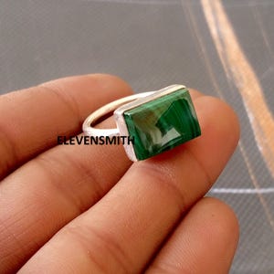 May include: A silver ring featuring a rectangular, vivid green malachite stone. The ring has a simple, contemporary design, highlighting the natural beauty of the gemstone. The word ELEVENSMITH is printed on the ring.